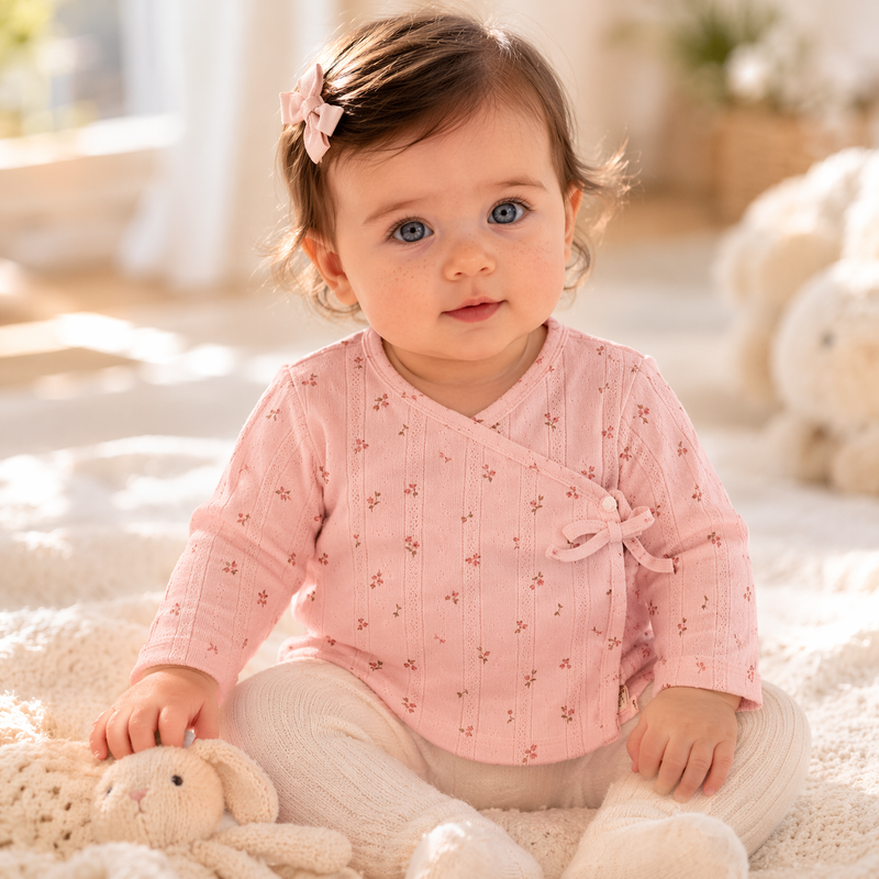Baby Mädchen (2–6 Monate) trägt ein rosa Wickelshirt mit feinem Blumenmuster, sitzt auf einer weichen Decke im warmen Tageslicht in einem hellen, gemütlichen Wohnraum.