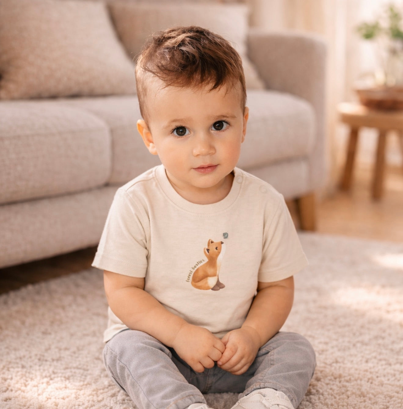 Kleiner Junge trägt ein beiges Kurzarm T Shirt mit Fuchs Motiv und sitzt auf einem weichen Teppich in einem gemütlichen Wohnzimmer