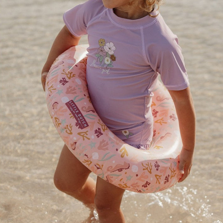 Kind trägt den Kinder Schwimmring Ocean Dreams Pink mit 50 cm Durchmesser am Strand. Der aufblasbare Schwimmring im maritimen Design ist ideal für Strand, Pool, Garten und sommerlichen Wasserspaß im Urlaub.